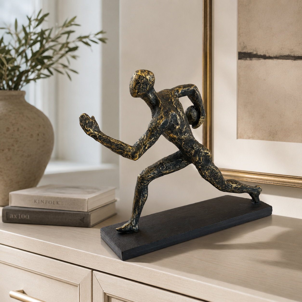 A bronze-finished running athlete sculpture with a textured metallic finish, displayed on a black rectangular base on a light wood console table.