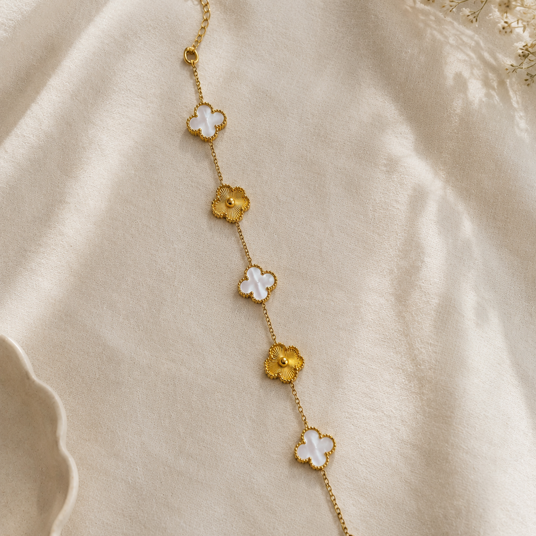 A delicate gold chain bracelet featuring alternating white and gold clover-shaped charms, laid out on a soft beige fabric background.