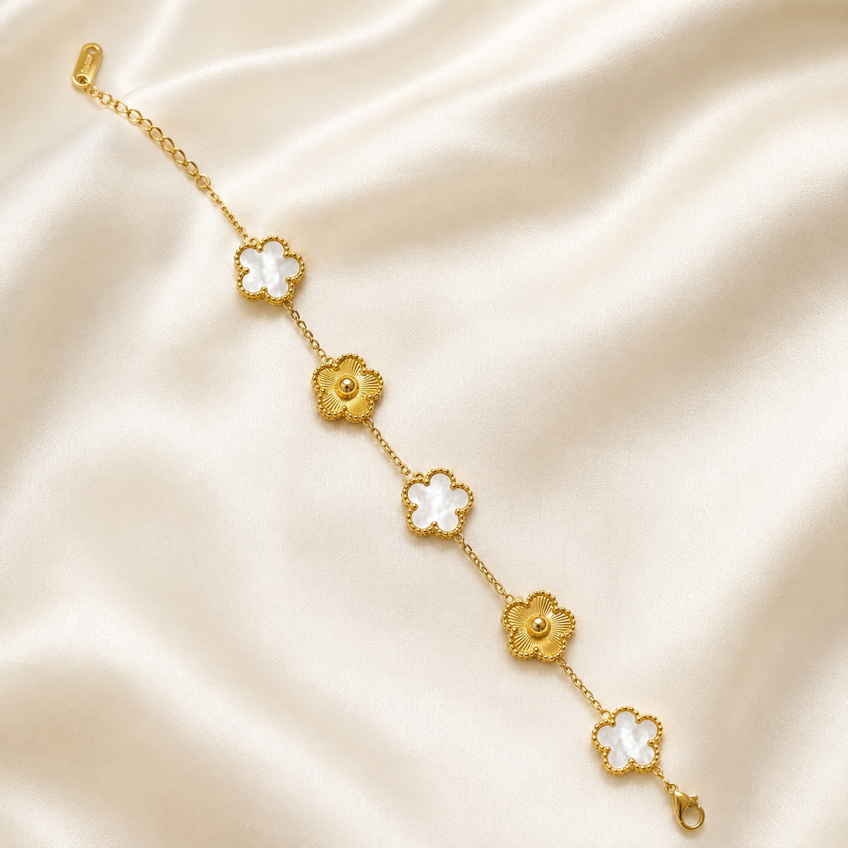 A gold chain bracelet featuring five alternating flower motifs, with three in gold and two in white mother-of-pearl, displayed on a draped cream satin fabric.