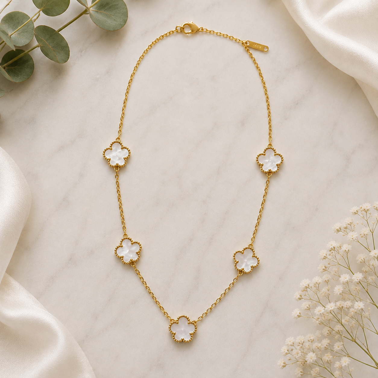 A gold-tone chain necklace with five alternating white and gold clover motifs, displayed on a light marble surface with decorative green leaves and white flowers.
