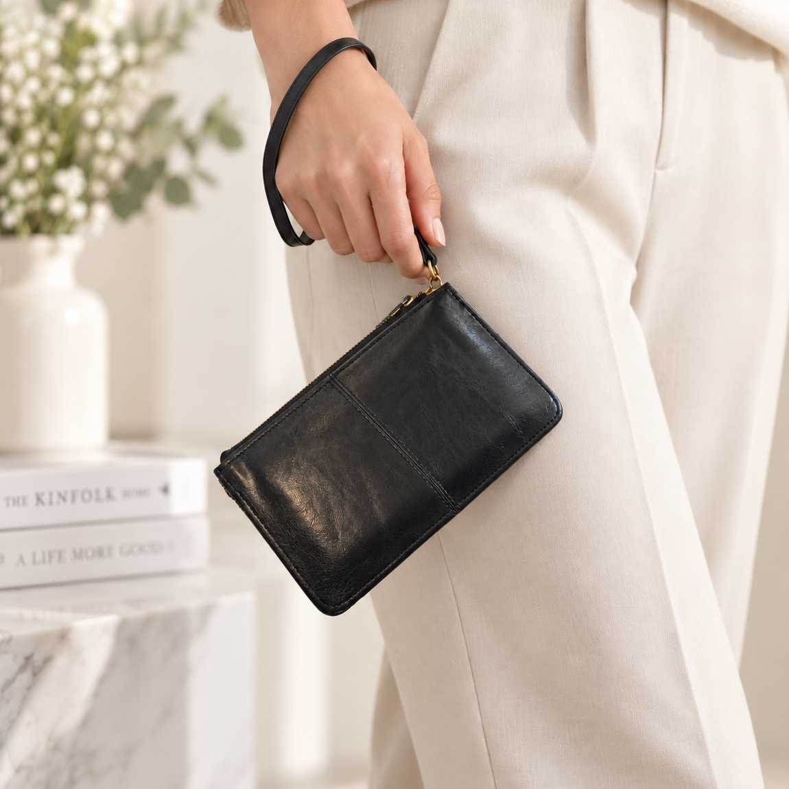A person wearing cream-colored knit pants holds a black leather wristlet pouch with a gold zipper in a bright, modern interior setting.