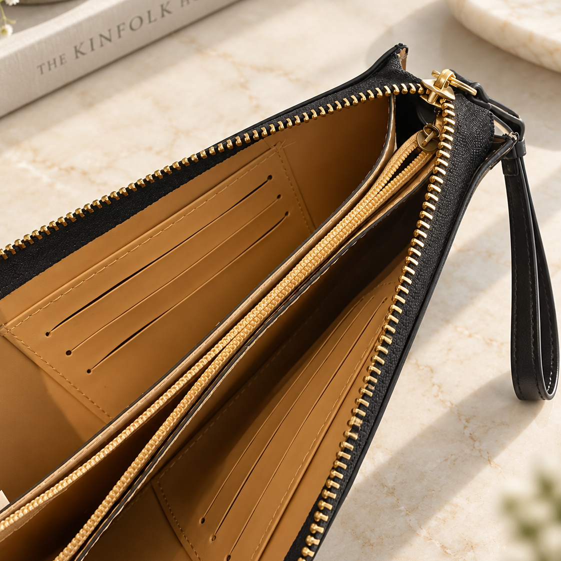 An open black leather clutch or wristlet with gold-tone zippers and a tan interior, photographed on a light marble surface with a book and stone props in the background.