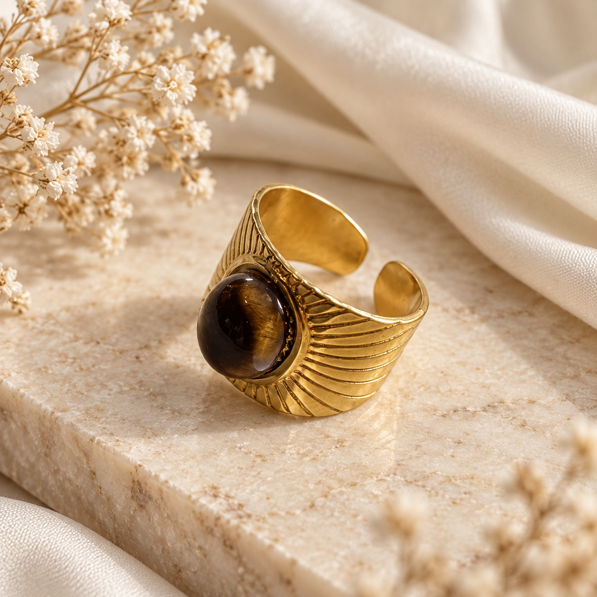 A gold-toned open cuff ring with a large round brown tiger's eye stone, styled on a beige stone slab with white fabric and dried flowers.