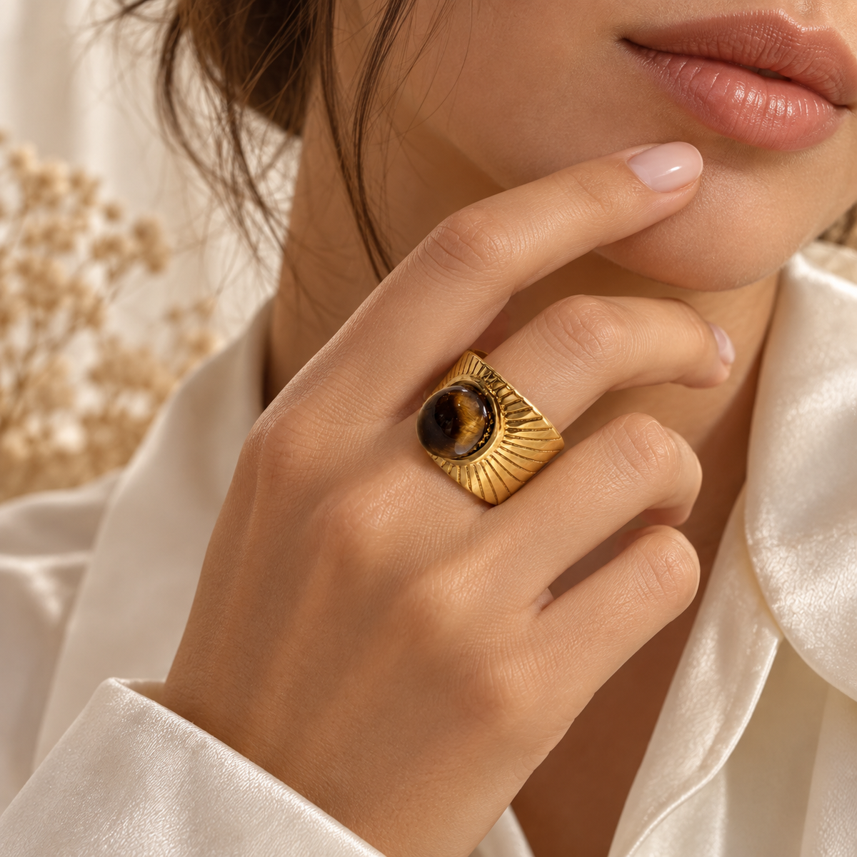 A close-up lifestyle shot of a model wearing a wide gold signet ring with a round brown tiger's eye stone, styled with a white satin shirt.