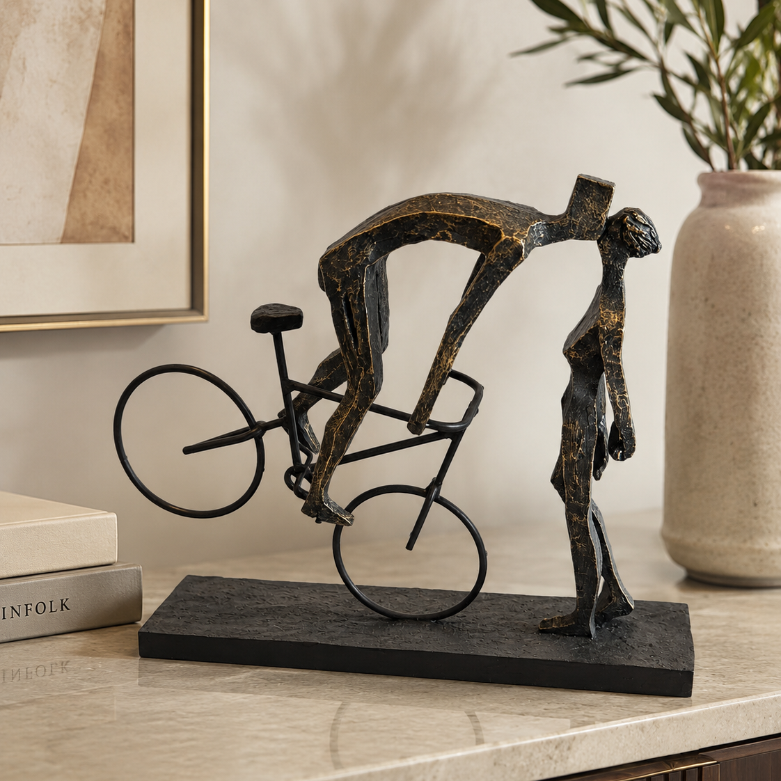 A bronze-finished decorative sculpture of two stylized human figures, one riding a bicycle and the other standing beside it, displayed on a black rectangular base on a marble countertop in a styled interior setting.