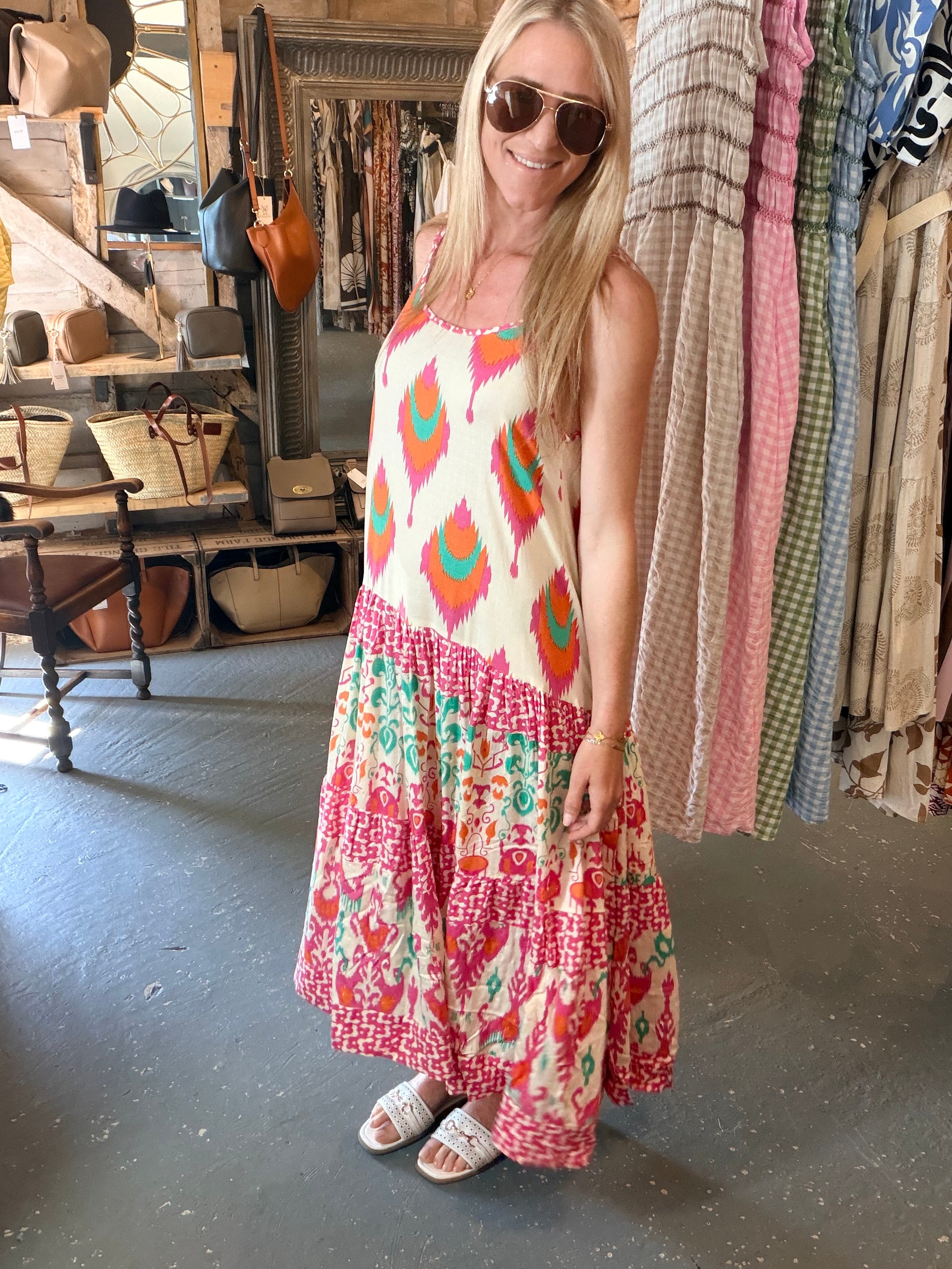 A woman wearing a sleeveless tiered maxi dress with a colorful ikat and floral print, standing inside a boutique shop.