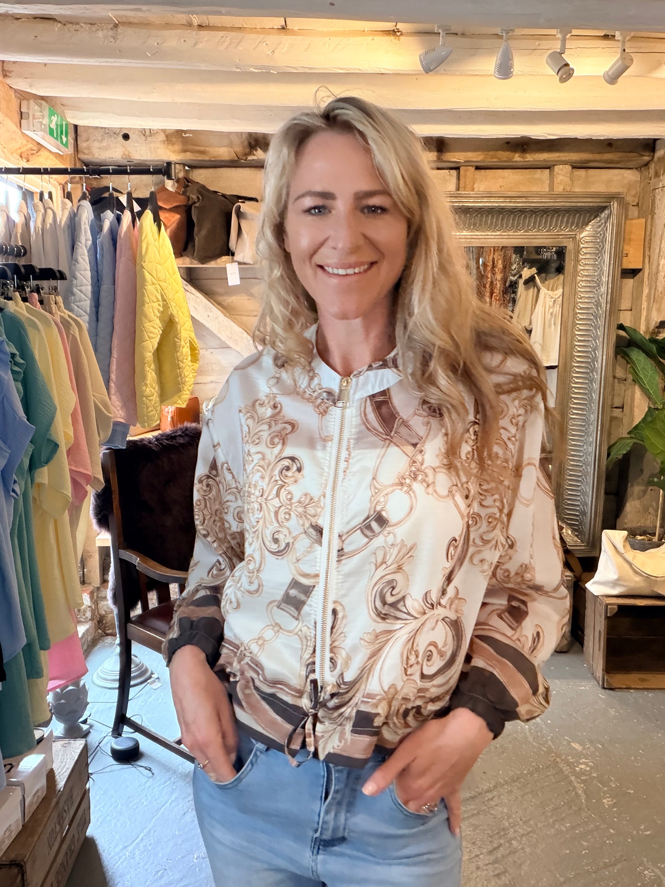 A woman with blonde hair stands inside a clothing boutique wearing a cream satin bomber jacket with a brown baroque scroll pattern, paired with light blue jeans.