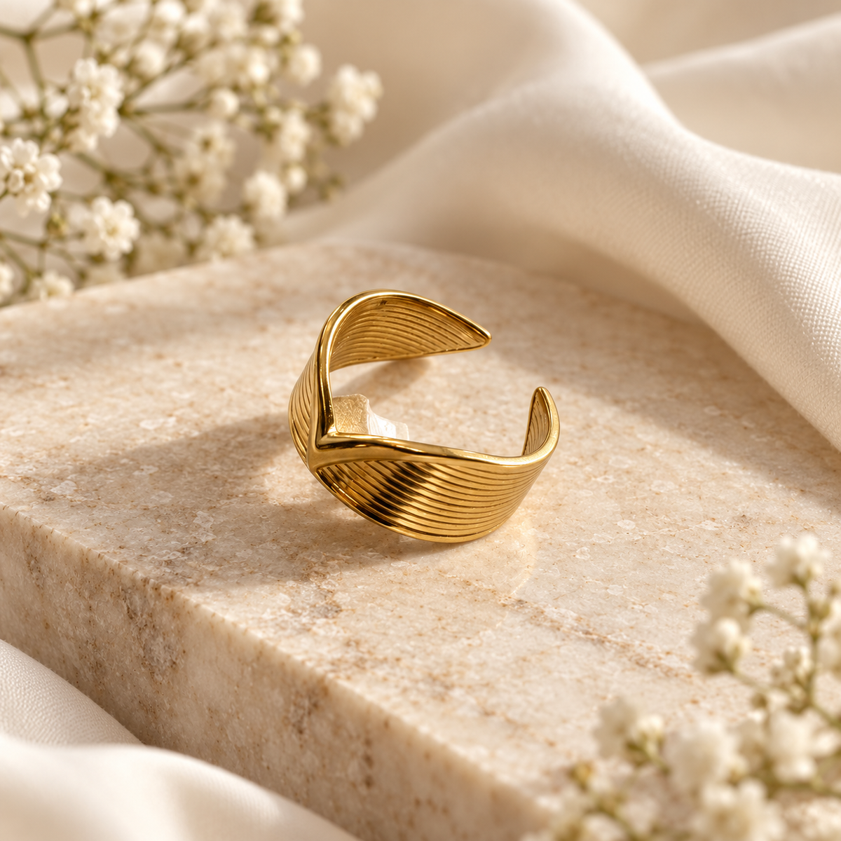 A gold-toned twisted ring with a ribbed texture, displayed on a beige stone slab with draped cream fabric and small white flowers.