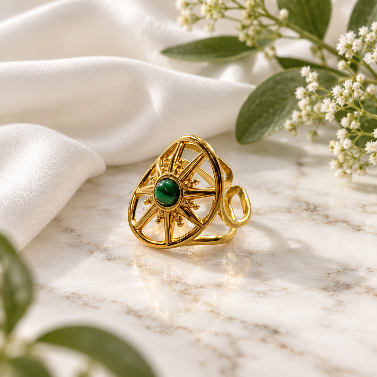 A gold-toned geometric ring with a central green oval stone, styled on a white marble surface with draped white fabric and small white flowers.