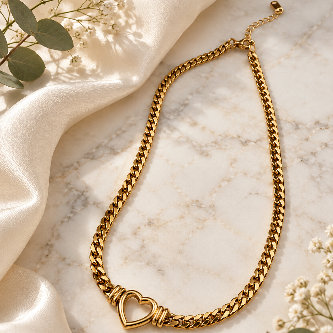 A gold-toned chain necklace with a heart-shaped pendant, styled on a white marble surface with cream satin fabric and small white flowers.