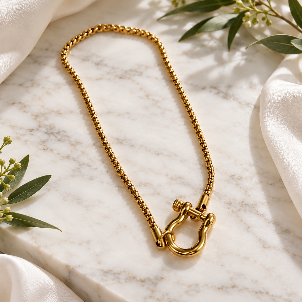 A gold-tone chain necklace with a large shackle clasp, styled on a white marble slab with white fabric and green leaves.