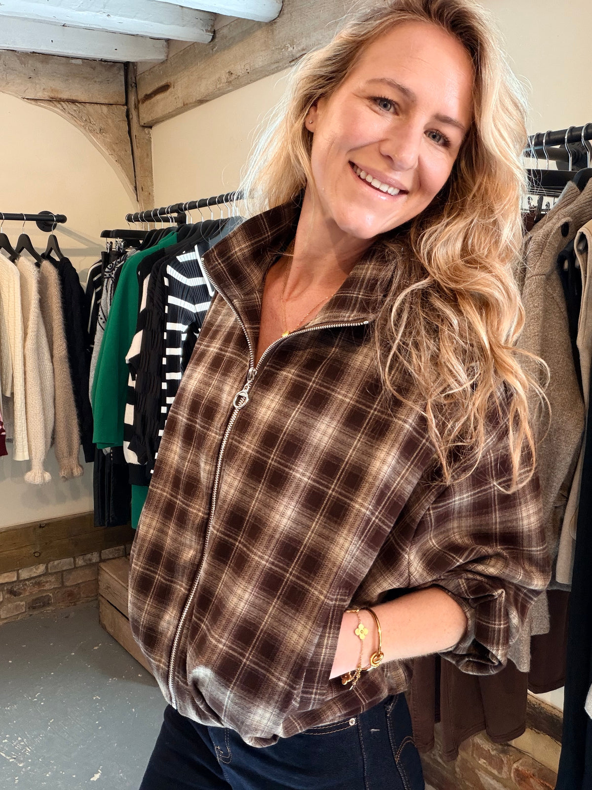 A smiling blonde woman wearing a brown and beige plaid zip-up fleece jacket, standing inside a clothing boutique with racks of garments in the background.