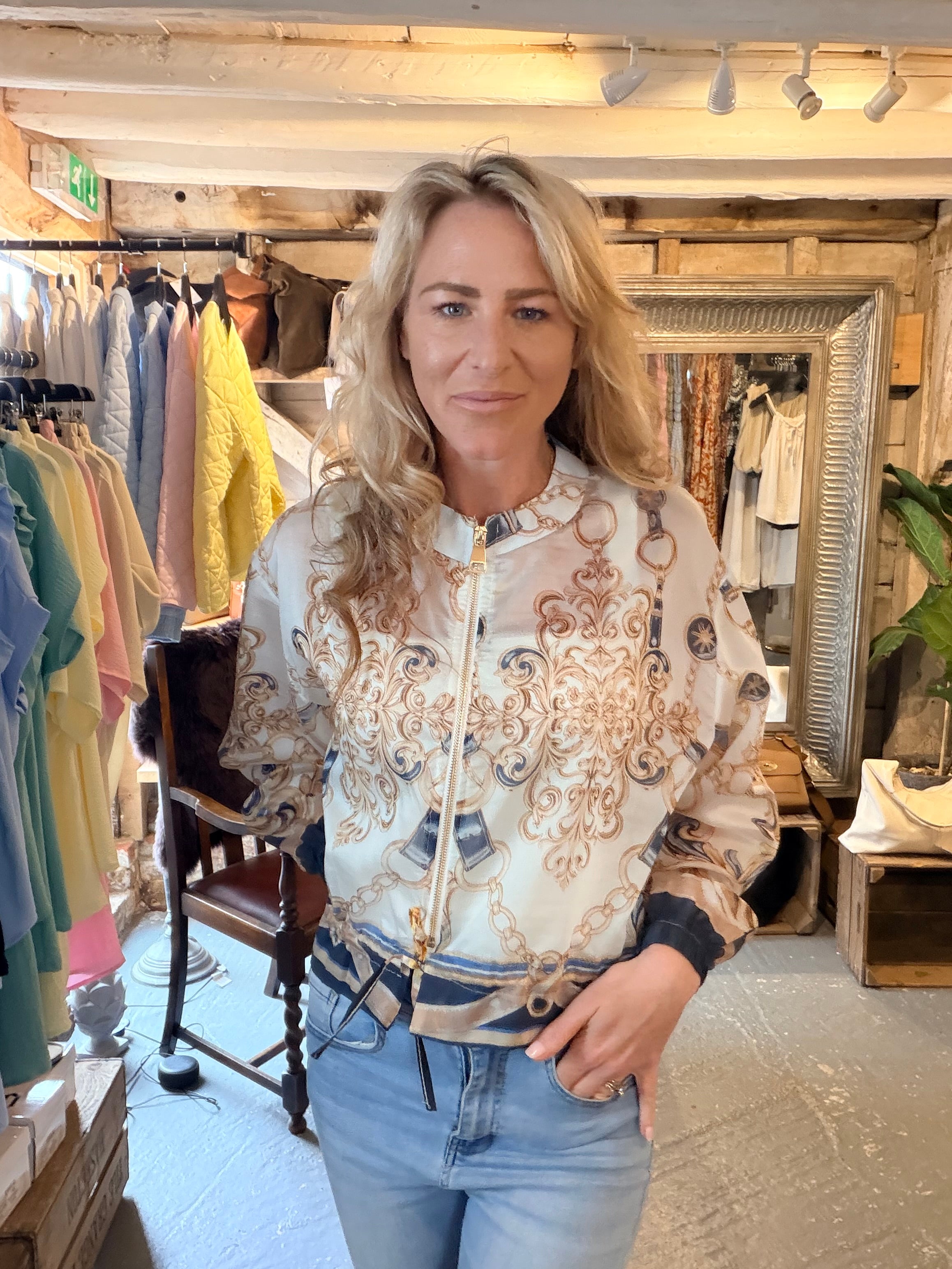 A woman with blonde hair stands in a clothing boutique wearing a cream-colored bomber jacket with an ornate gold and brown baroque-style pattern, paired with light blue jeans.