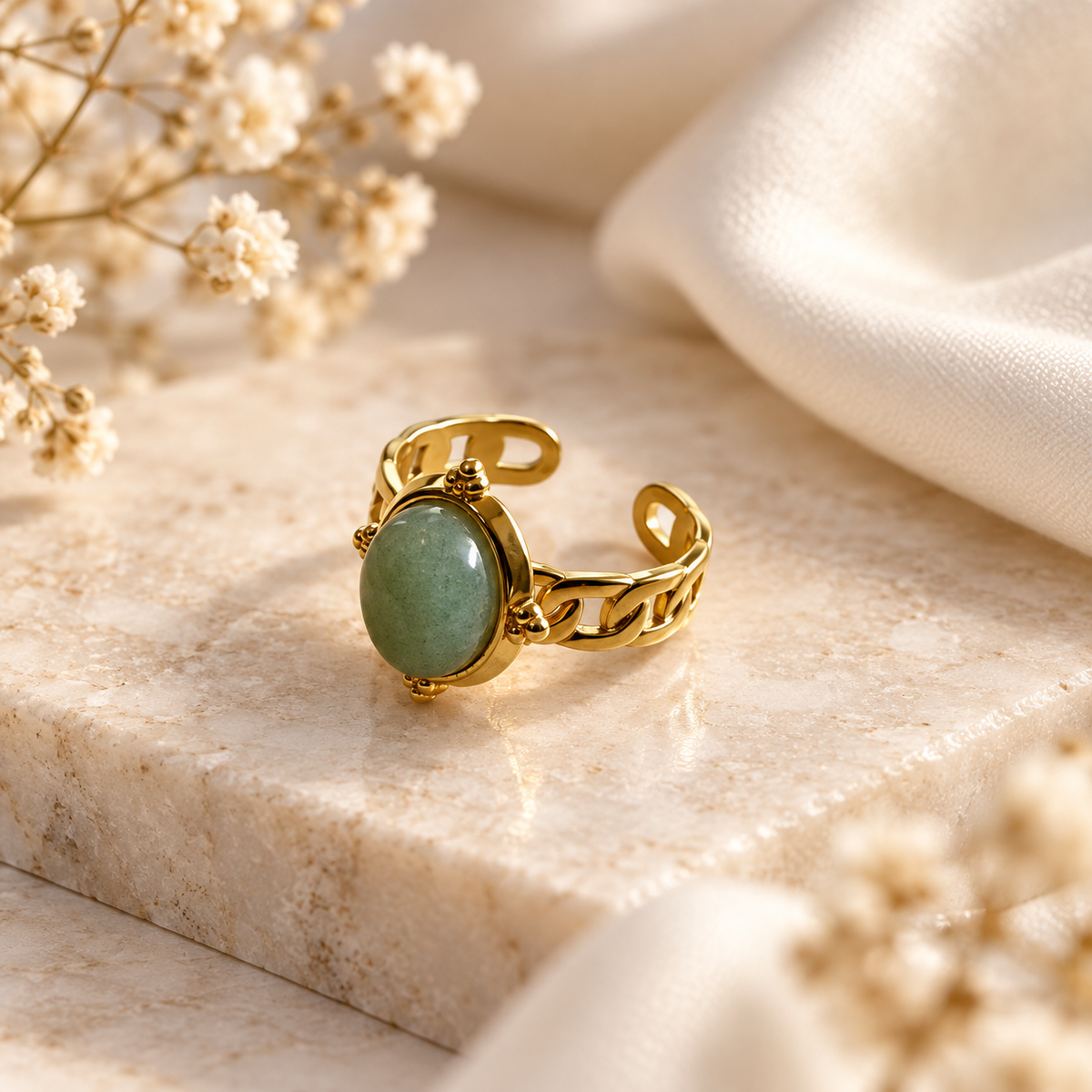 A gold-toned adjustable ring with an oval light green stone, styled on a beige marble block with cream fabric and dried flowers.