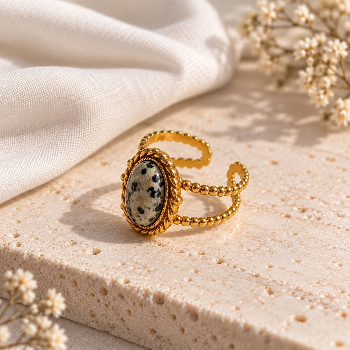 A gold-toned open ring bangle featuring an oval Dalmatian-spotted stone in a rope-like bezel setting, styled on a textured beige stone slab with draped white fabric and dried flowers.