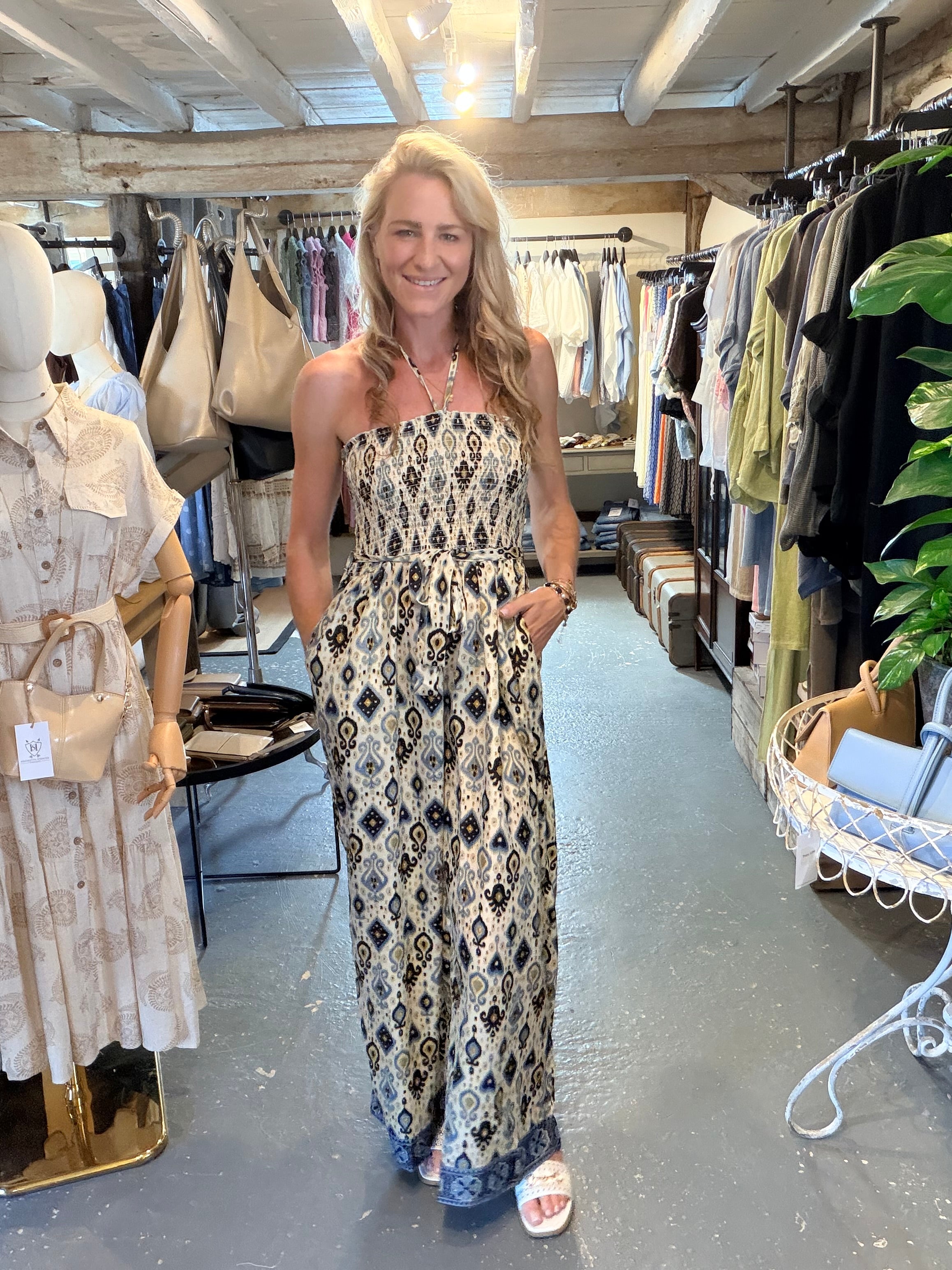 A woman stands in a clothing boutique wearing a strapless jumpsuit with a black, blue, and beige geometric pattern. The boutique has clothing racks, mannequins, and a gray concrete floor.