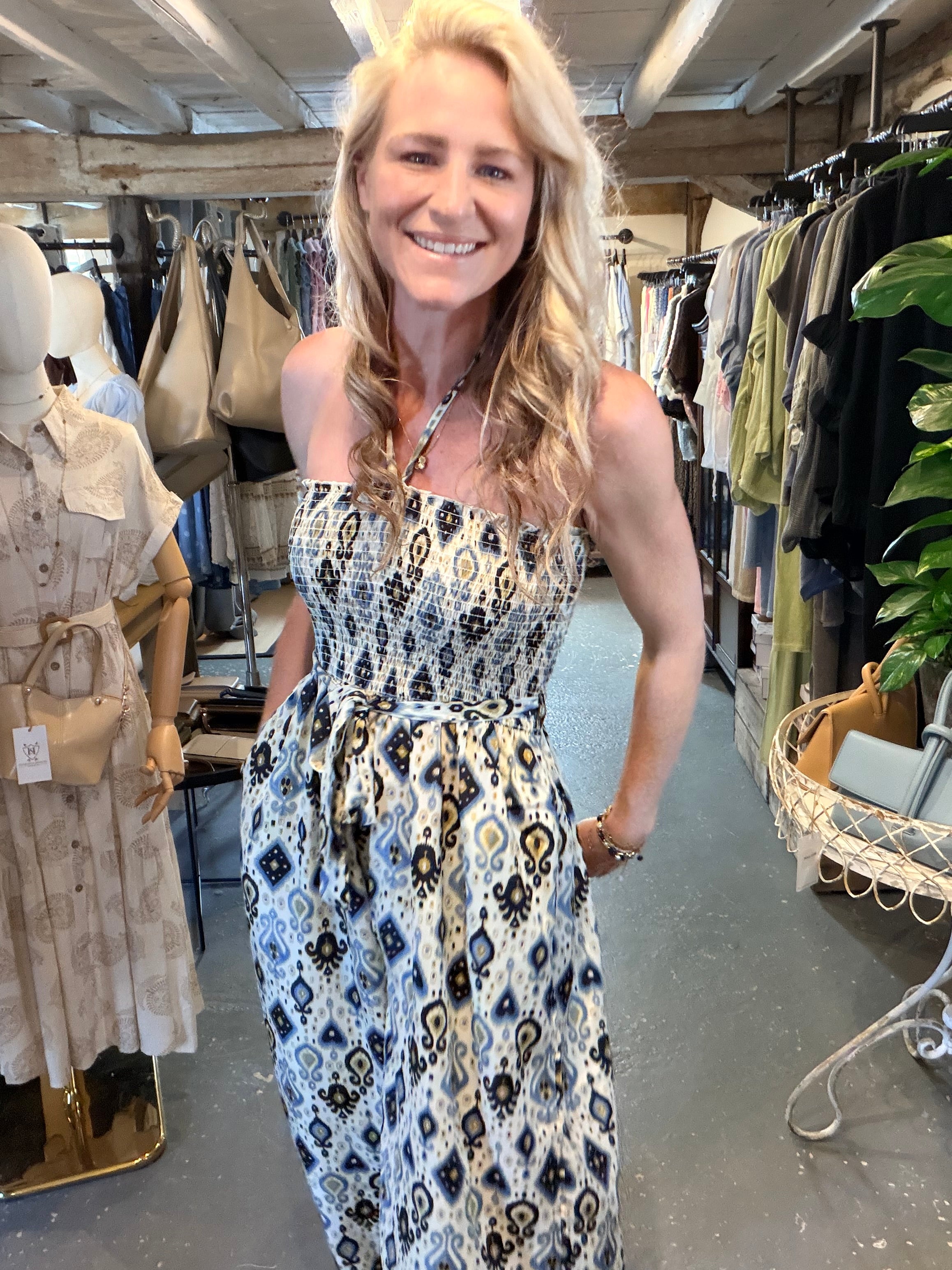 A smiling blonde woman wearing a strapless midi dress with a black, white, and blue ikat pattern, standing inside a clothing boutique.