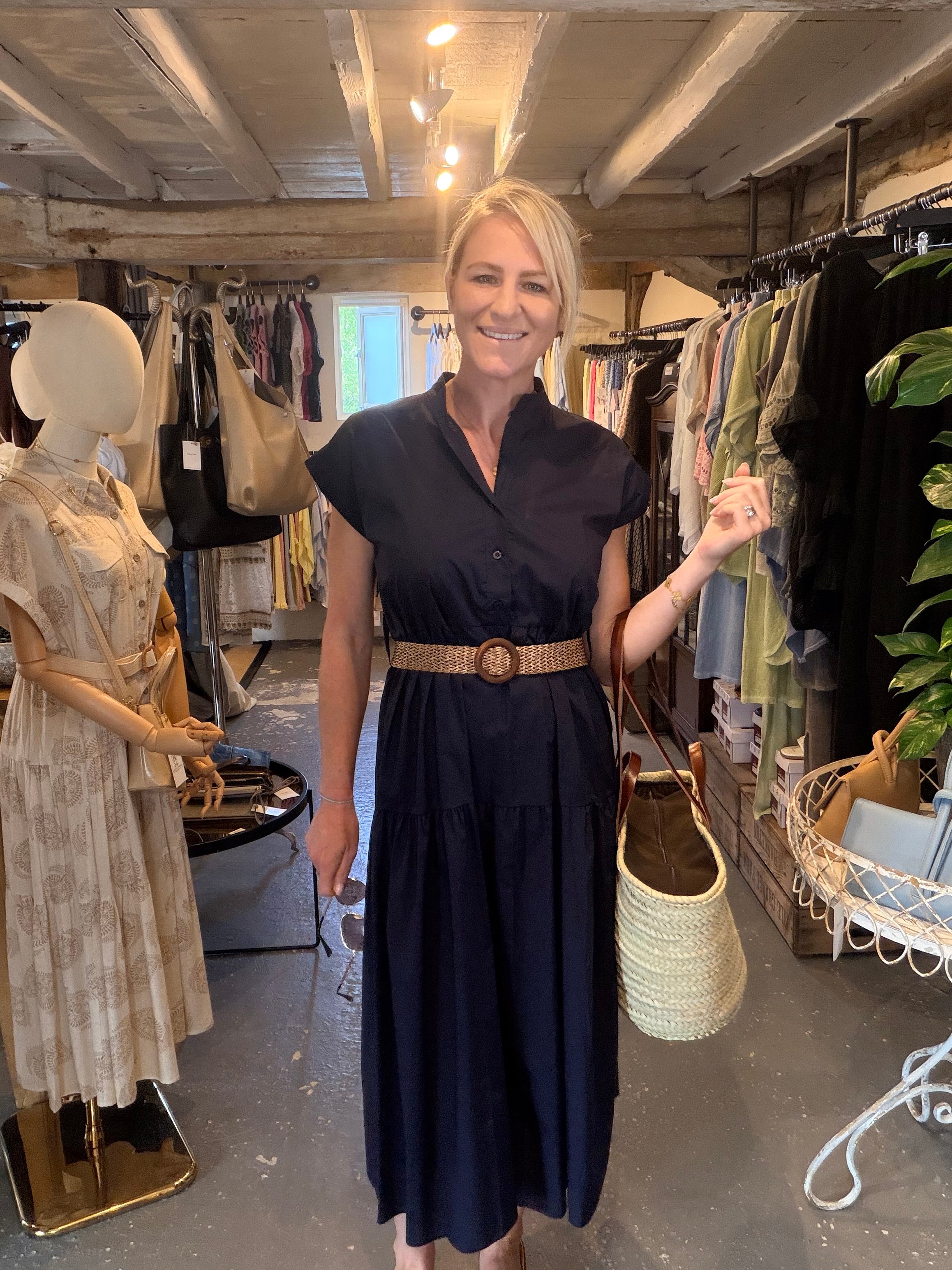 A woman stands inside a clothing boutique wearing a dark navy midi dress with a wide tan belt and holding a woven straw bag. Mannequins and racks of clothes are visible in the background.