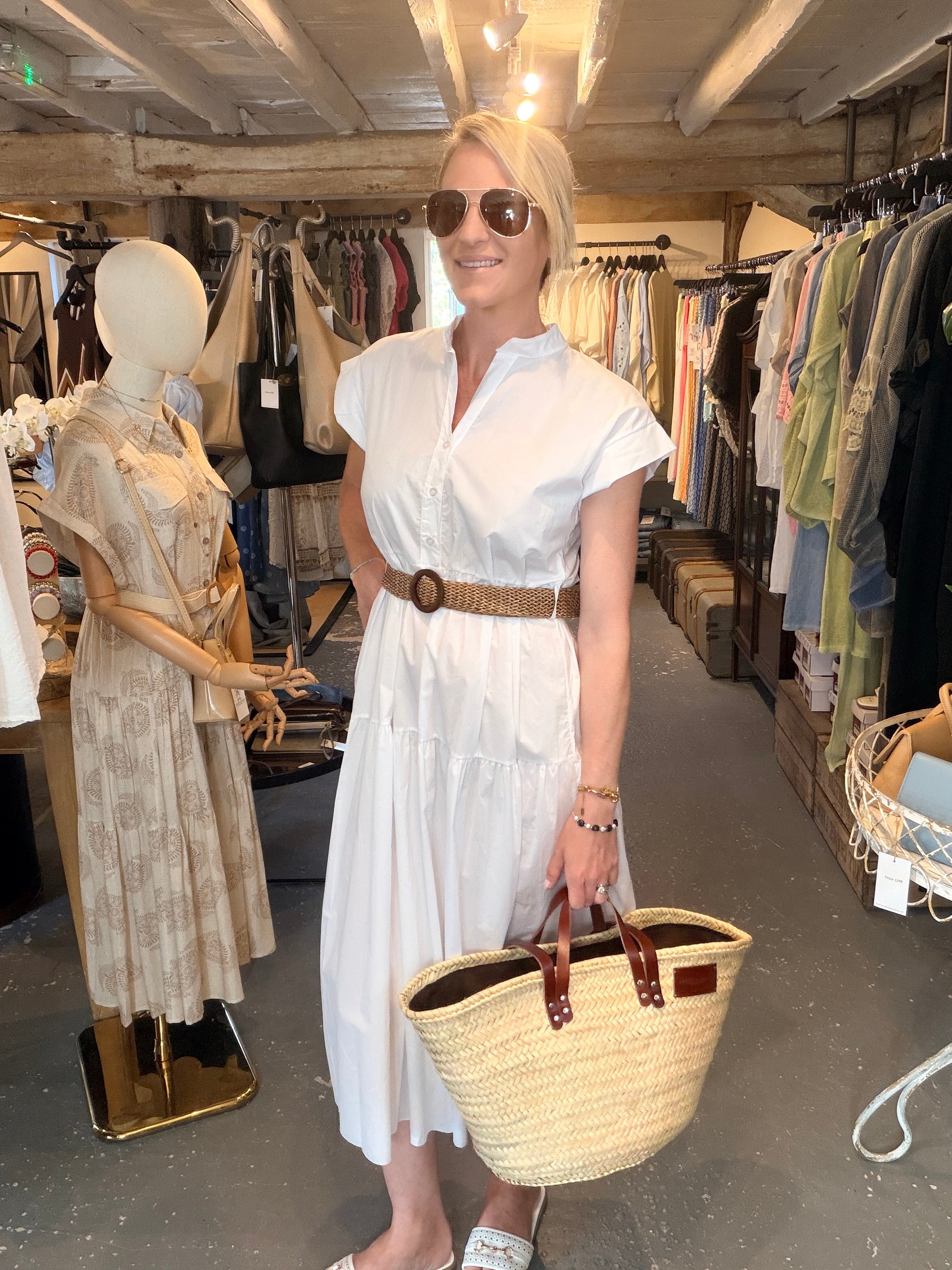 A woman wearing a white short-sleeve midi shirt dress with a brown belt, sunglasses, and holding a woven straw tote bag with brown leather handles, standing inside a clothing boutique.
