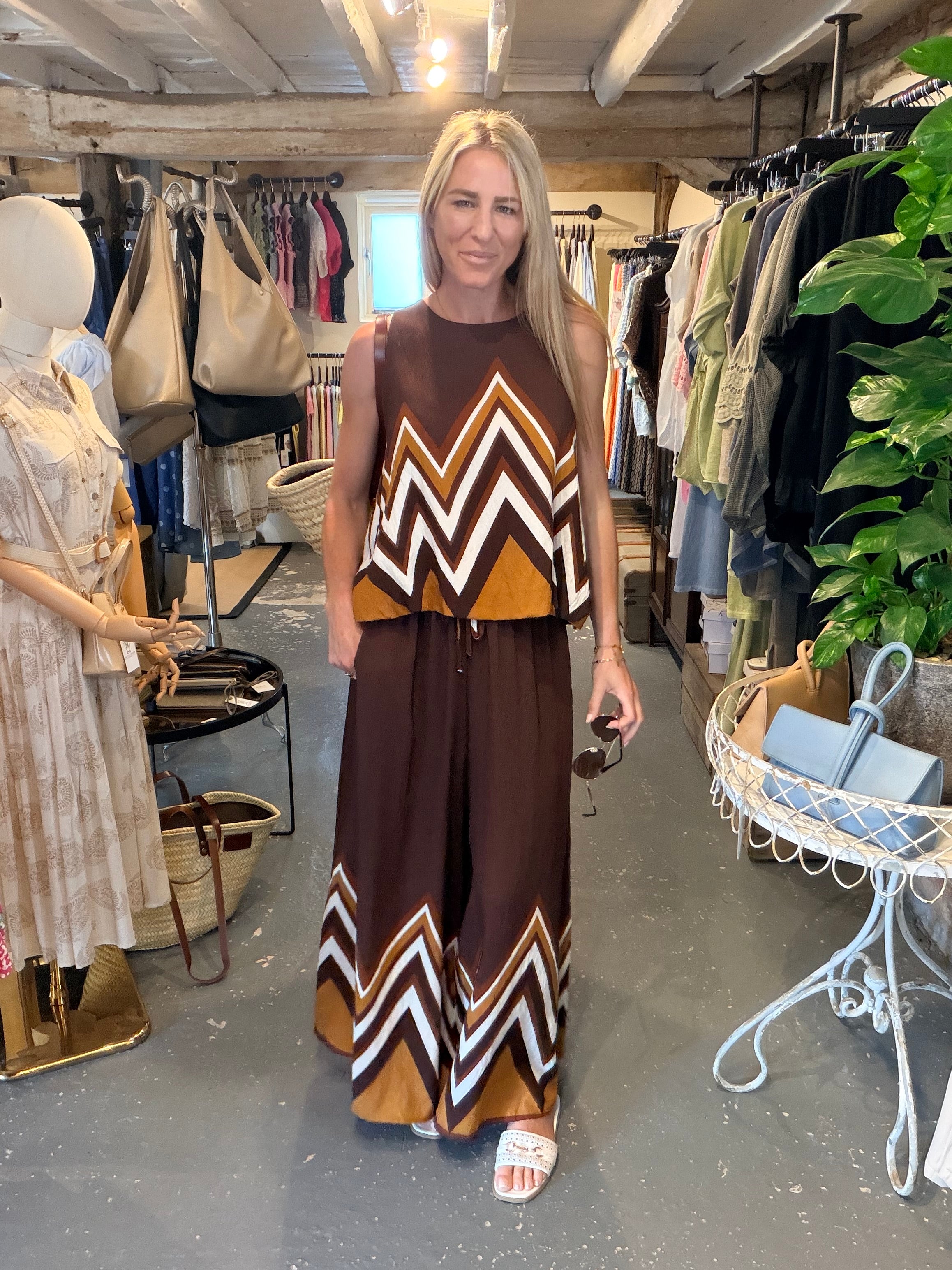 A woman modeling a brown sleeveless jumpsuit with a brown, beige, and white chevron pattern, standing inside a clothing boutique.