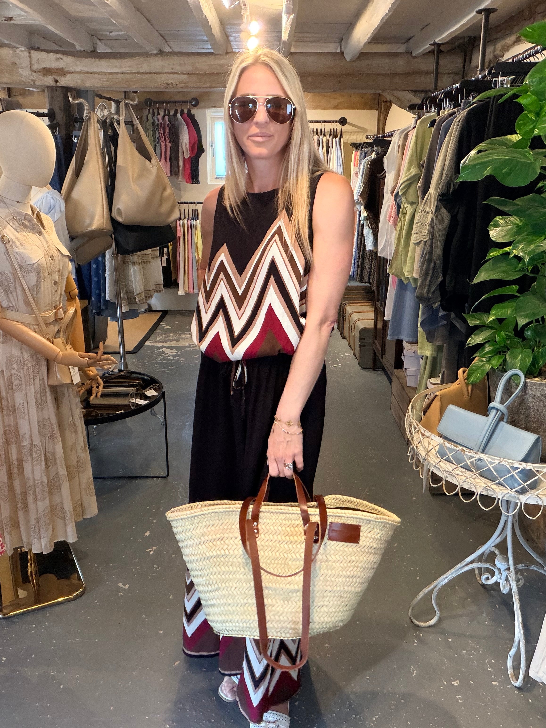 A woman wearing sunglasses and a sleeveless chevron top stands inside a boutique holding a large woven straw tote bag with brown leather handles.