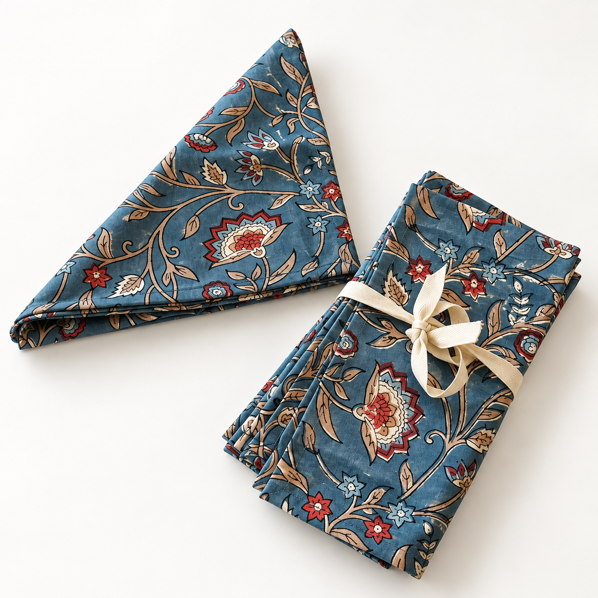 A set of folded patterned napkins in a dusty teal-blue color, featuring a traditional floral and vine print in red, beige, and white. One napkin is unfolded to display the full pattern, while the other is folded and tied with a cream ribbon.