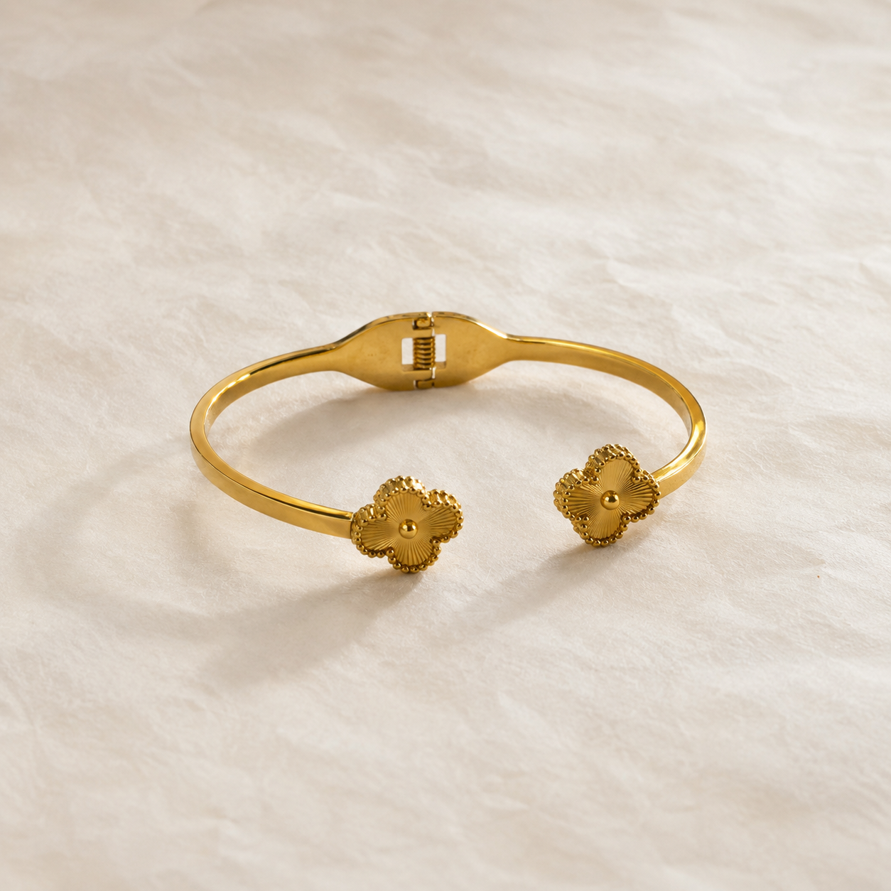 A gold-tone cuff bracelet with two small flower-shaped end details, photographed on a textured off-white fabric background.