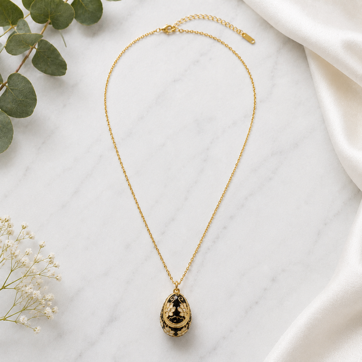A gold chain necklace with a teardrop-shaped pendant featuring black and gold floral detailing, displayed on a white marble surface with green leaves and white fabric.