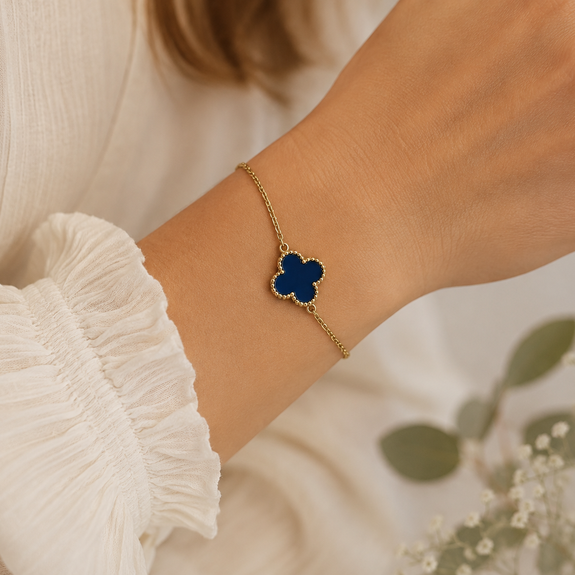 A gold-toned chain bracelet with a dark blue four-leaf clover pendant, worn on a woman's wrist. The person is wearing a white ruffled blouse, with soft-focus green leaves in the background.
