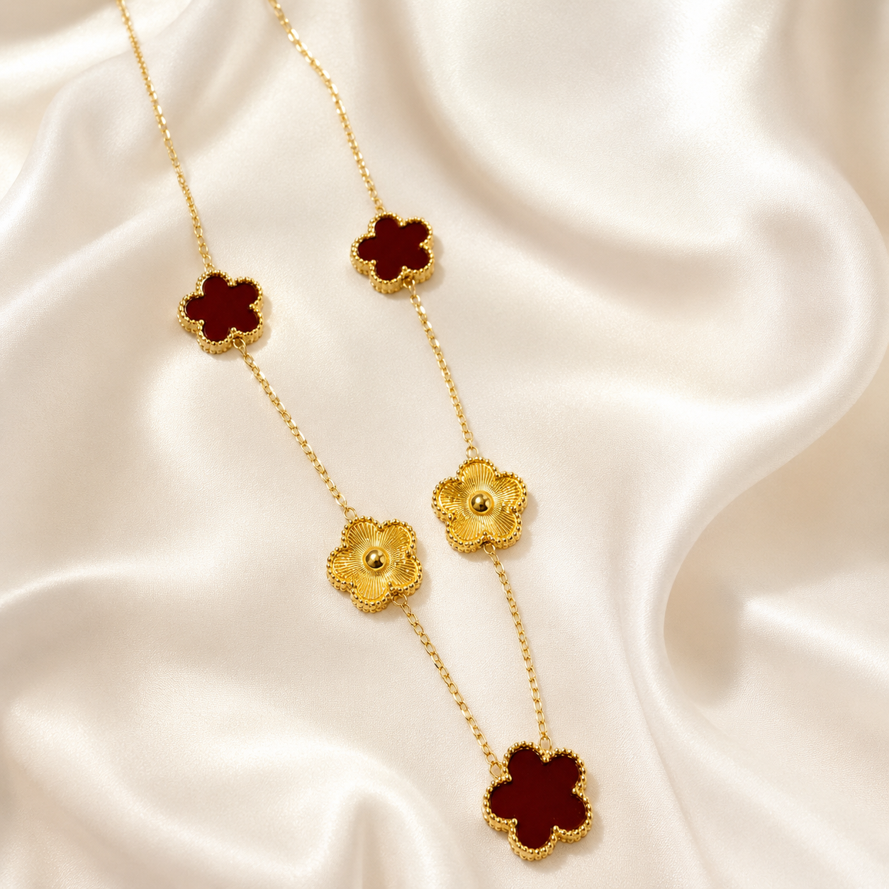 A gold chain necklace with four clover-shaped motifs, featuring deep red inlay and textured gold borders, displayed on a draped white satin fabric.