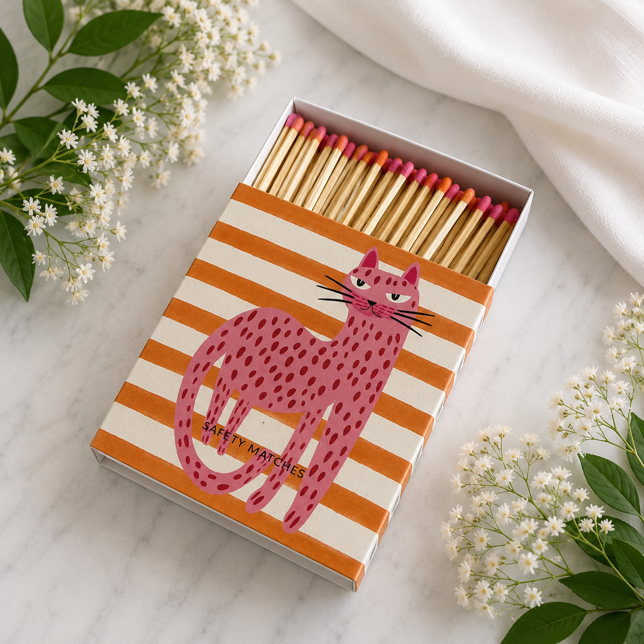 A matchbox filled with wooden matches, styled on a light marble surface with white flowers and a white fabric. The box lid features orange and cream stripes and a pink spotted cat illustration with the text 'SAFETY MATCHES'.
