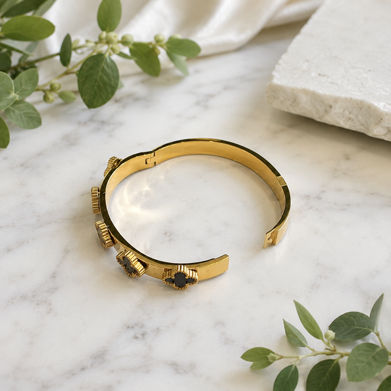 A gold cuff bracelet with textured gear-like details and a central dark blue gemstone, styled on a white marble surface with green leaves and a white stone slab.