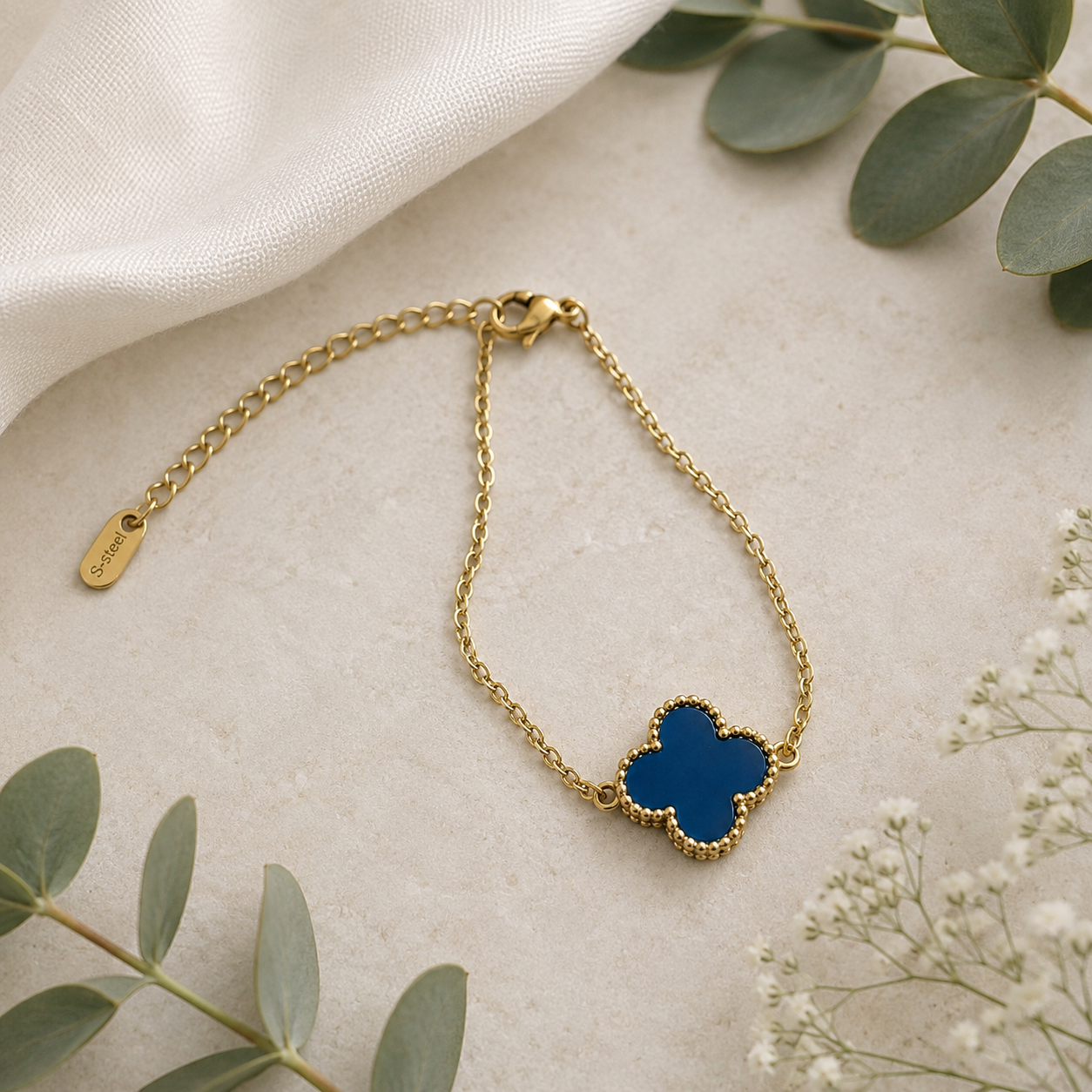 A gold chain bracelet with a blue clover-shaped pendant, styled on a light textured surface with green leaves and white fabric.