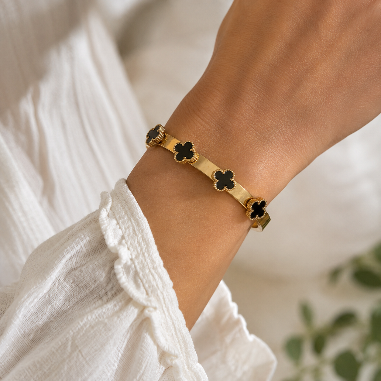 A gold-toned bracelet with four black clover motifs, worn on a person's wrist over a white textured blouse.