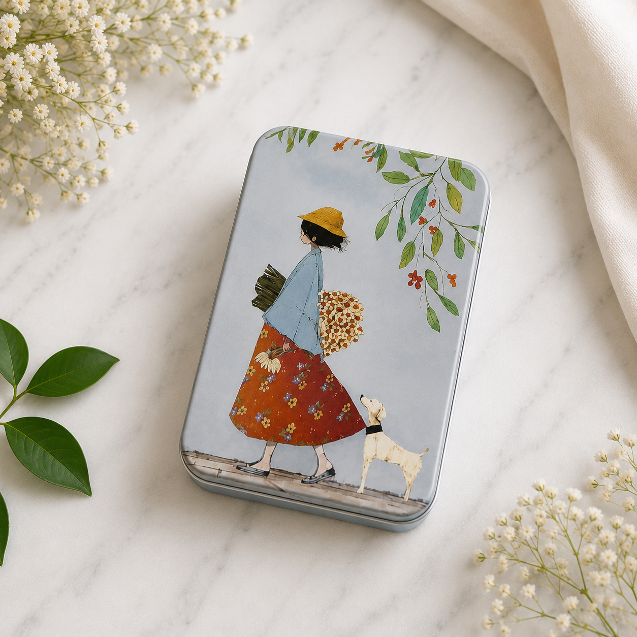 A rectangular metal tin with rounded corners, decorated with an illustration of a woman in a yellow hat and floral skirt walking with a white dog, placed on a white marble surface with baby's breath flowers and a cream fabric around it.