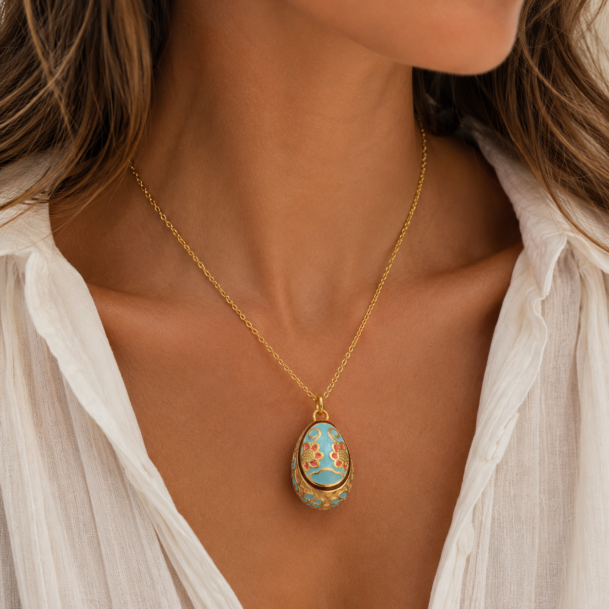 A gold chain necklace with an egg-shaped pendant featuring a light blue enamel background and orange floral patterns, worn by a model in a white blouse.