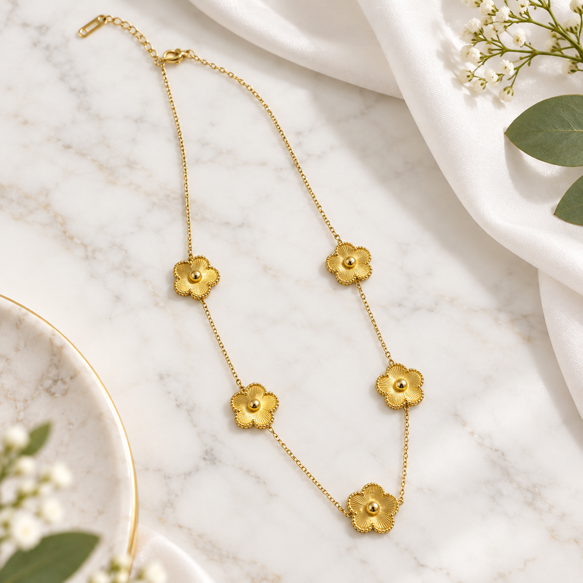 A delicate gold chain necklace featuring five textured flower charms, styled on a white marble surface with white fabric and green leaves.