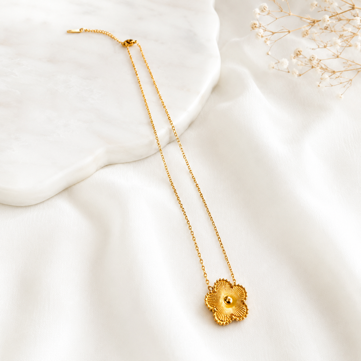 A delicate gold chain necklace with a textured, flower-shaped pendant, styled on a soft white fabric background with a white marble slab and dried baby's breath.