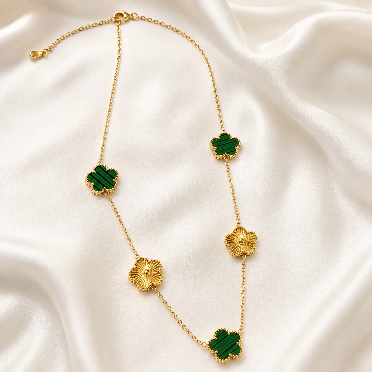 A gold chain necklace featuring five alternating clover-shaped charms, three of which are dark green and two are gold, displayed on a draped white satin fabric.