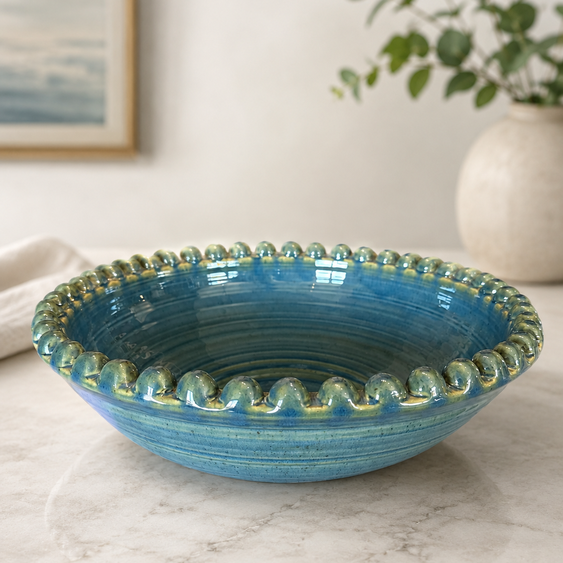 A round ceramic bowl with a glossy blue and green glazed finish and a wavy rim, styled on a marble surface with a blurred plant in the background.