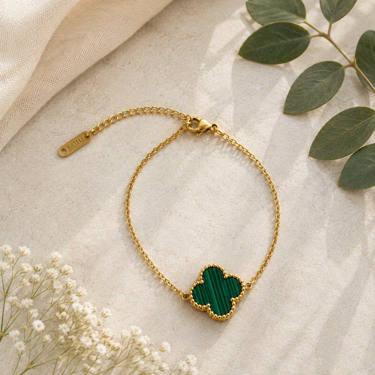 A gold chain bracelet with a four-leaf clover pendant featuring a textured green inlay, styled on a beige fabric background with green leaves and white baby's breath flowers.