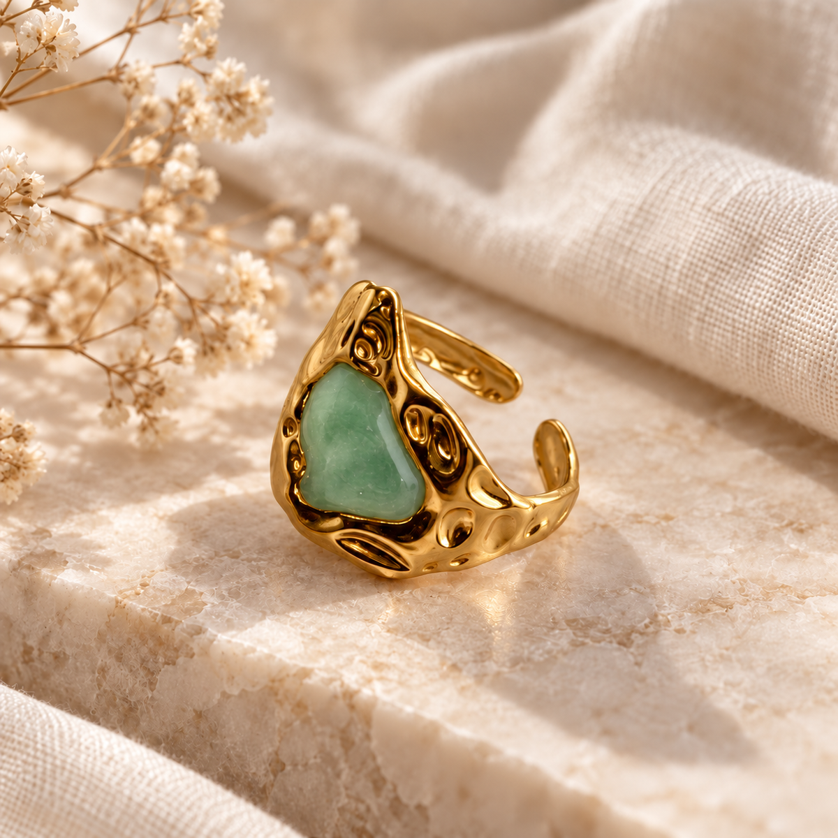 A gold-toned open ring with a heart-shaped light green stone, styled on a textured beige stone surface with cream fabric and dried flowers.