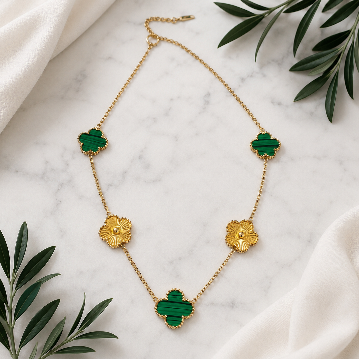 A gold chain necklace with five green and gold Alhambra-inspired clover and flower charms, styled on a white marble surface with white fabric and green leaves.