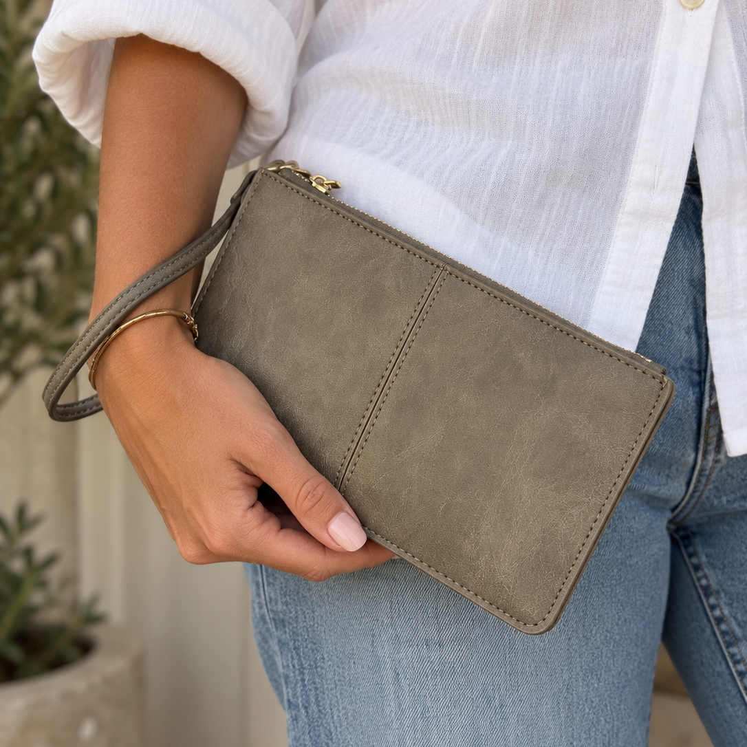 A person wearing a white button-up shirt and light blue jeans holds a taupe faux-leather clutch with a matching wristlet strap and gold-tone zipper.