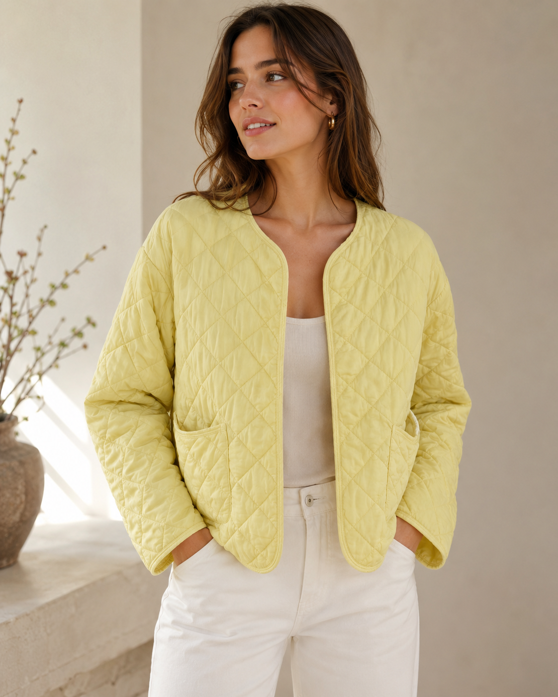 A woman wearing a light yellow quilted jacket over a white top and white pants, standing in a softly lit indoor setting with a neutral wall and a vase of branches in the background.