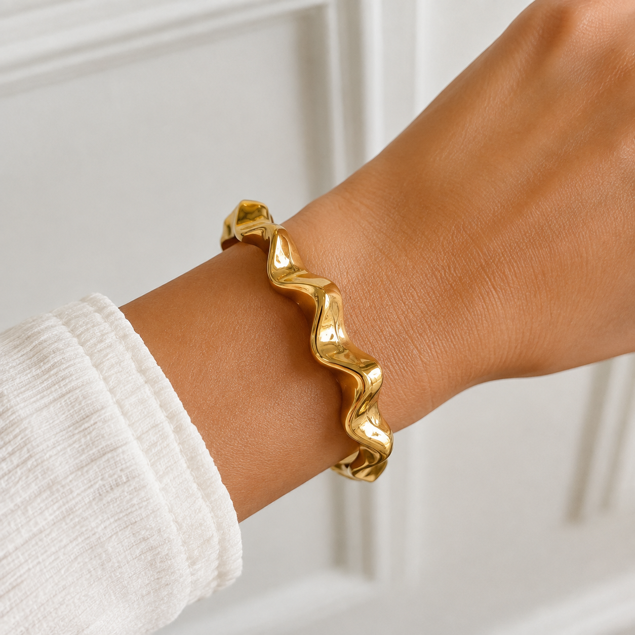 A polished gold wavy bangle bracelet worn on a person's wrist, paired with a white ribbed long-sleeve top, set against a soft-focus white paneled background.