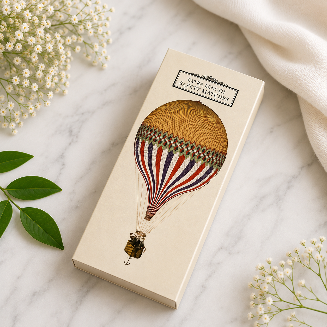 A cream-colored rectangular box of 'Extra Length Safety Matches' featuring an illustration of a hot air balloon, styled on a white marble surface with baby's breath flowers and a white fabric napkin.
