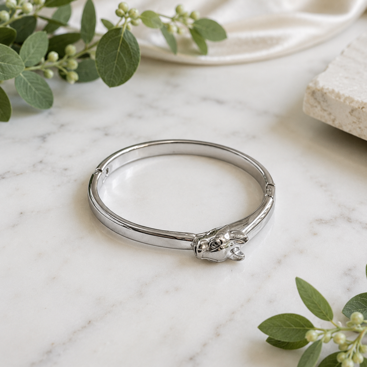 A polished silver-toned bangle bracelet with a sculpted animal head at the front, displayed on a white marble surface with green leaves and neutral props in the background.