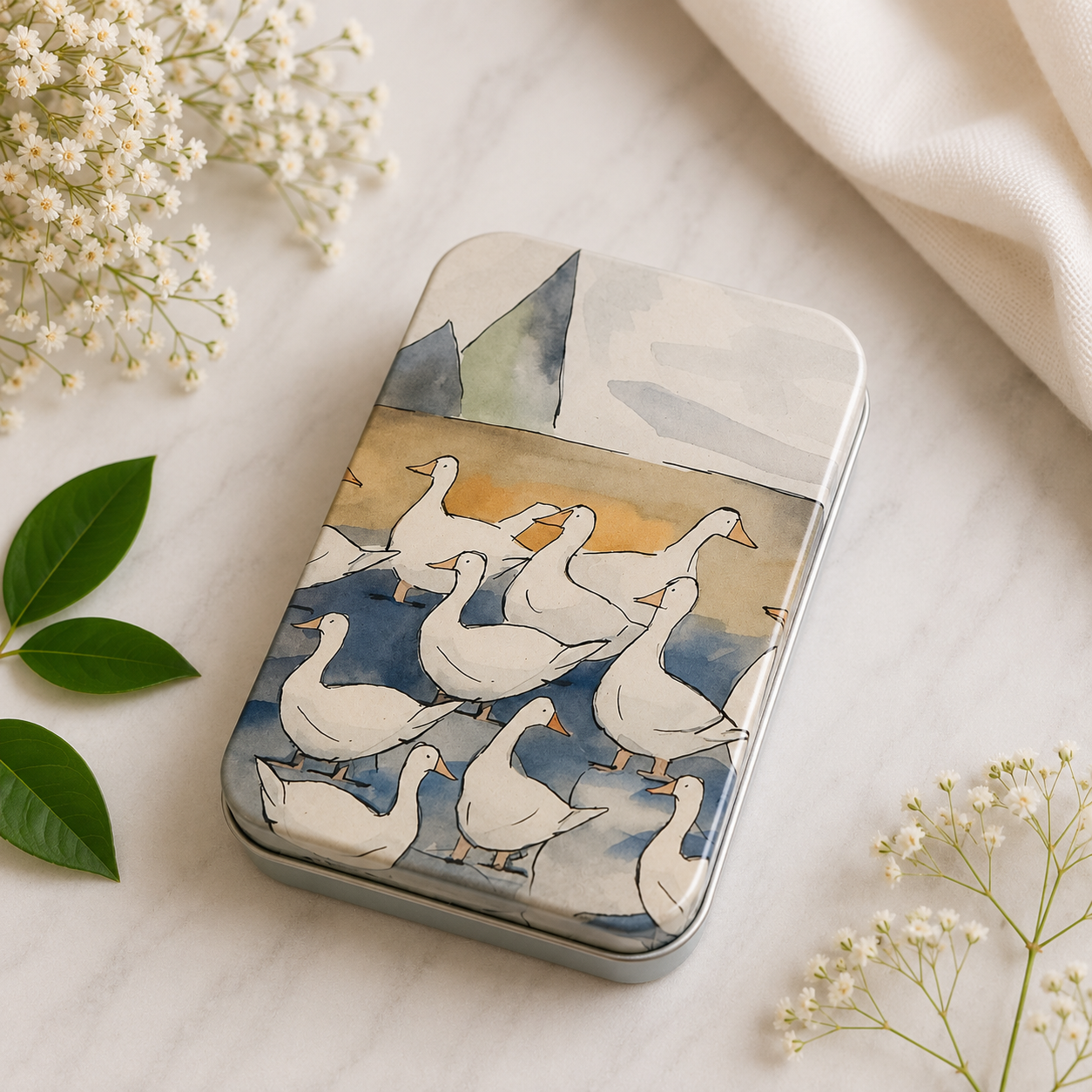 A rectangular metal tin with rounded corners, decorated with an illustration of a flock of white ducks on a blue and beige background. The tin is styled on a light marble surface with white baby's breath flowers and green leaves.