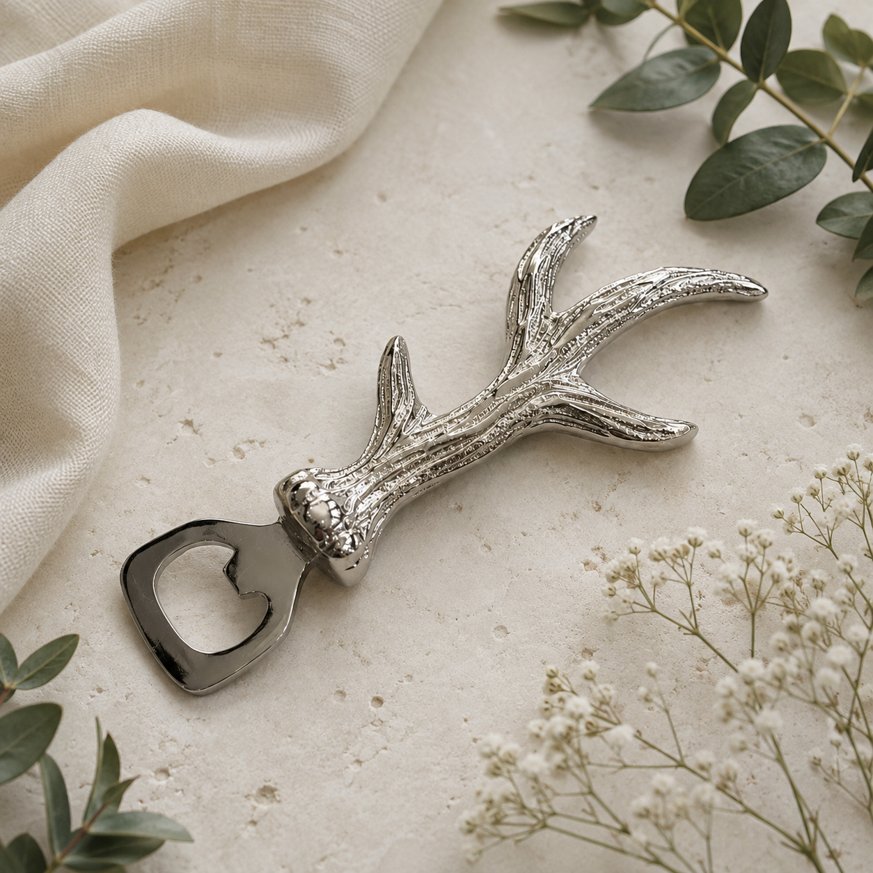 A silver deer antler bottle opener styled on a textured beige surface with a cream fabric napkin, green eucalyptus leaves, and small white baby's breath flowers.
